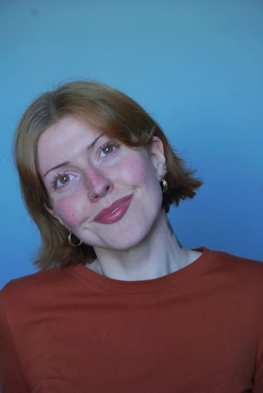 A woman with short light auburn hair, hazel eyes, and multiple earrings, wearing a burnt orange top, looking slightly upward with a gentle smile against a blue background. Shot by Emma Burton Studio.