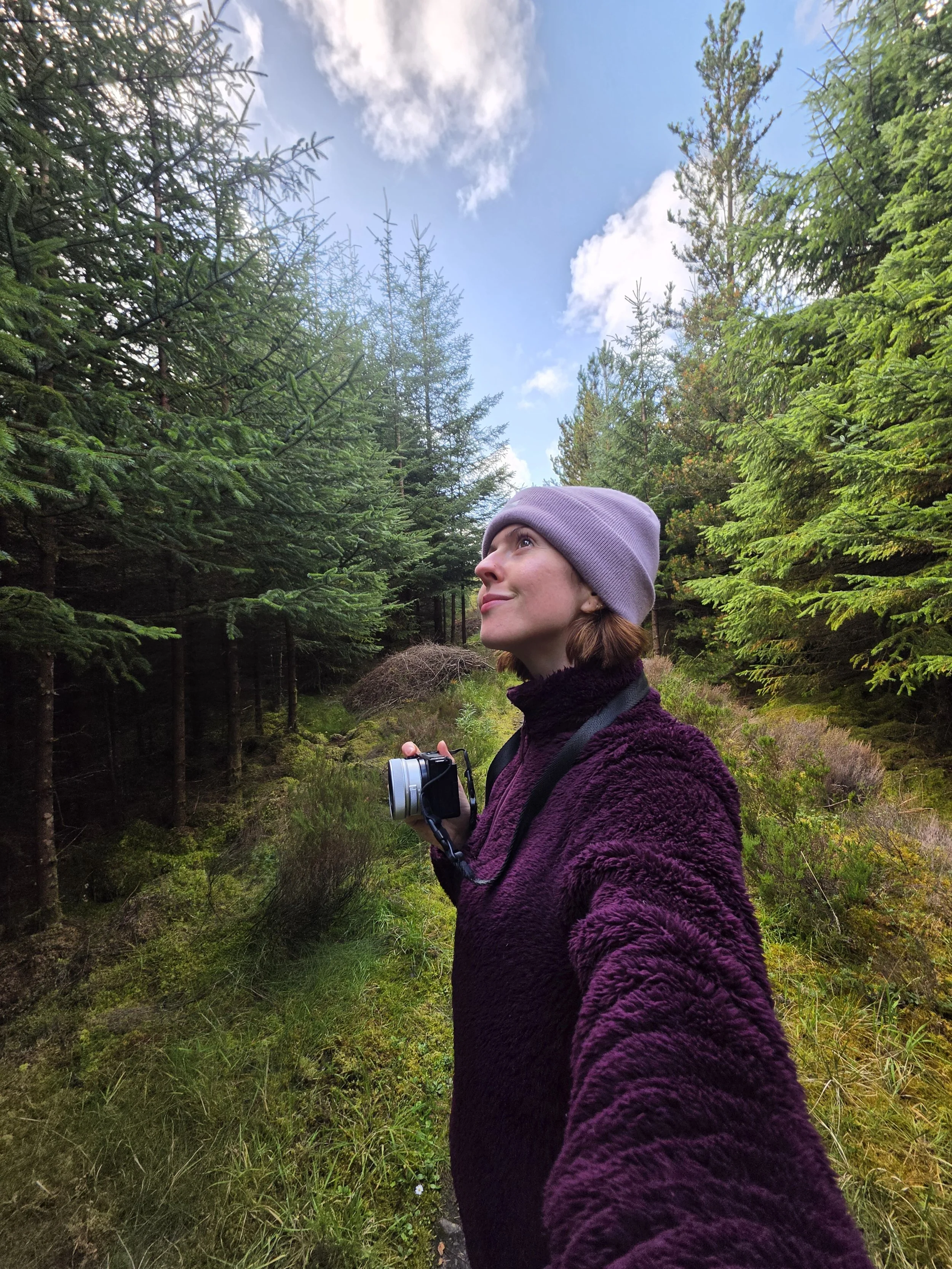 A woman with short auburn hair wearing a lilac beanie and purple fleece is standing in a lush green forest in the Scottish Highlands, holding a camera in her right hand, looking upward with a slight smile at the trees and sky.
