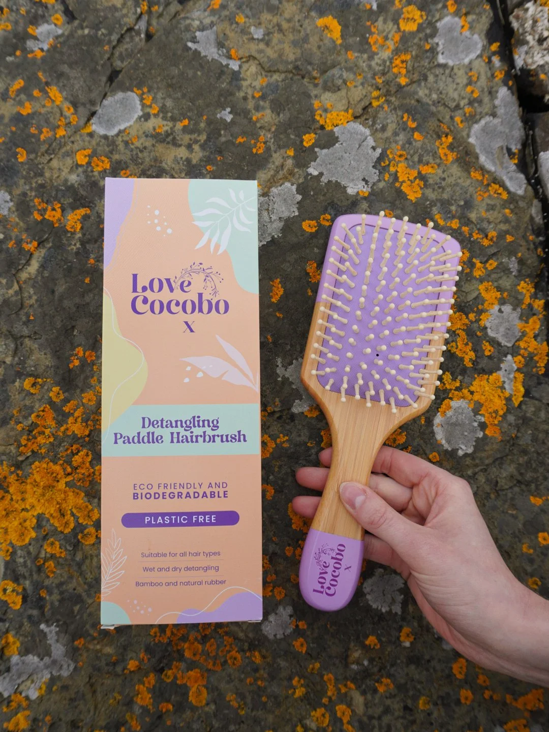 Hand holding a Love Cocobo eco-friendly bamboo hairbrush next to its packaging on textured dark rock with orange lichen in Scottish Highlands, shot by Emma Burton Studio