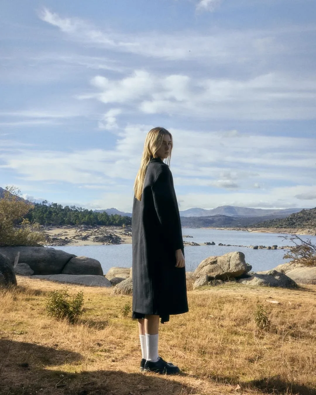 retrato mujer rubia en lago, cielo azul con nubes, chica joven, pelo largo, foto pantano, madrid,  revista moda, gabardina negra, otoño, calcetas blancas con zapatos negros, fashion, nostalgia, melancolía, soledad, cabello rubio, editorial