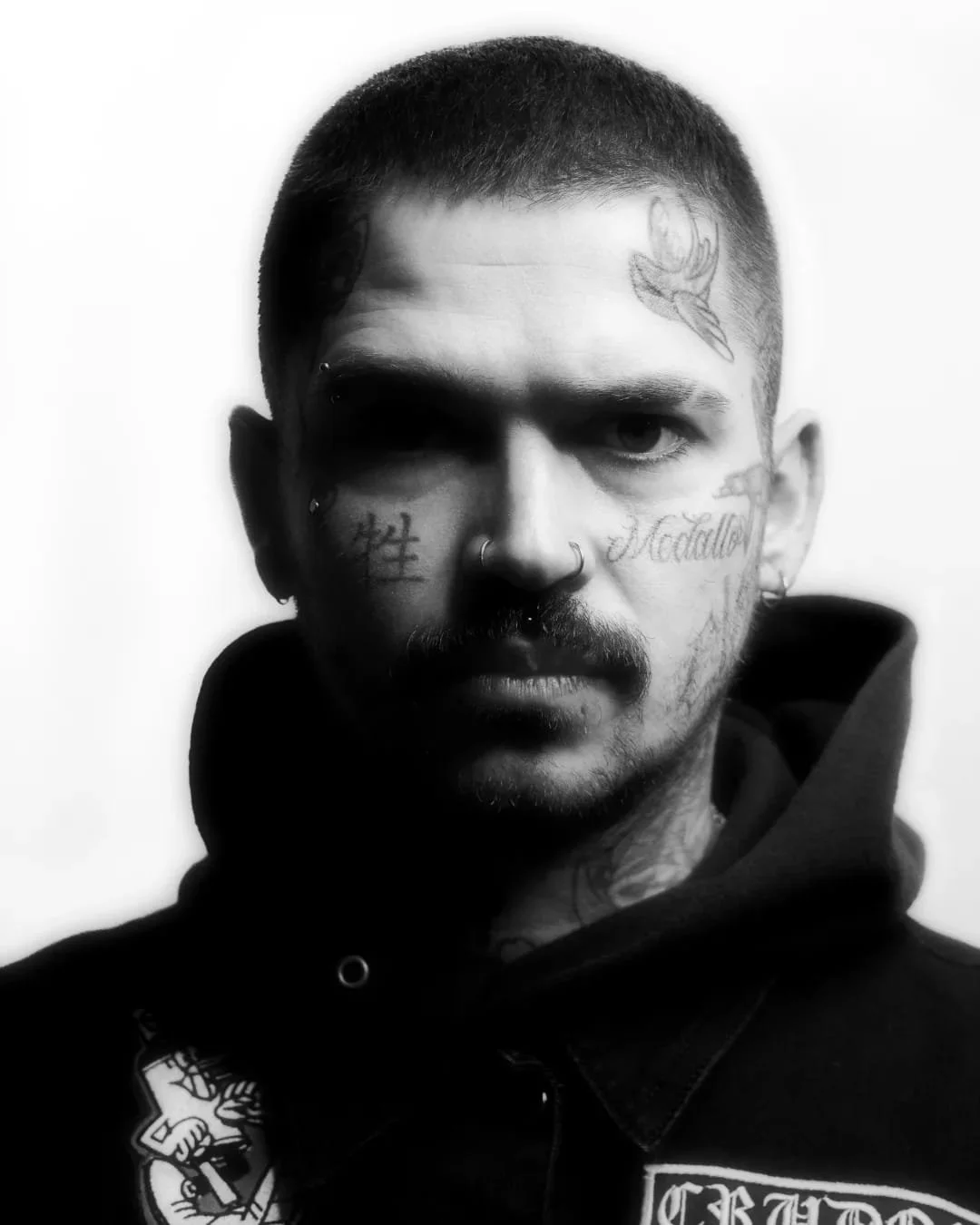 rudo, colombiano, foto chico, hombre tatuado, punk, alternativo, retrato blanco y negro, tatuajes, tattoo, parches, crudos, sudadera negra, chaqueta, chamarra negra, rapado, medallo, pelo corto, cara tatuada, madrid retrato, perforaciones, piercings