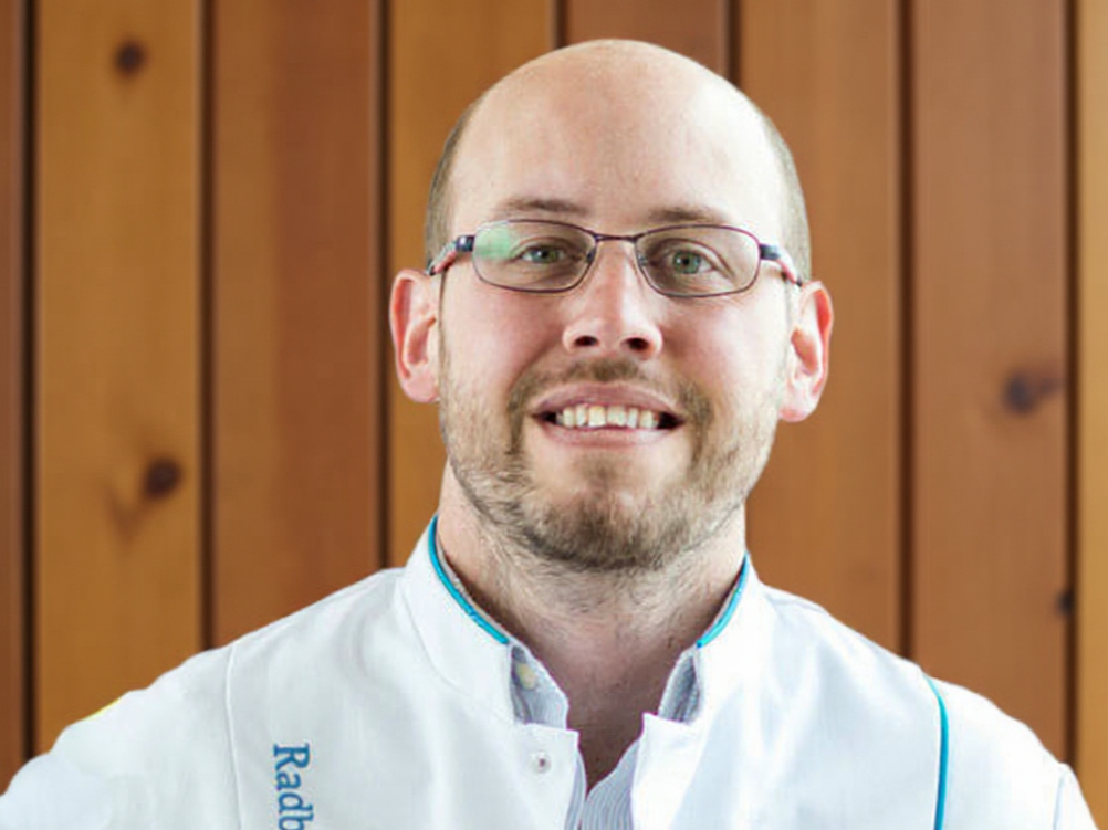 A man wearing glasses and a white chef coat with blue trim, standing in front of a wooden wall, smiling.