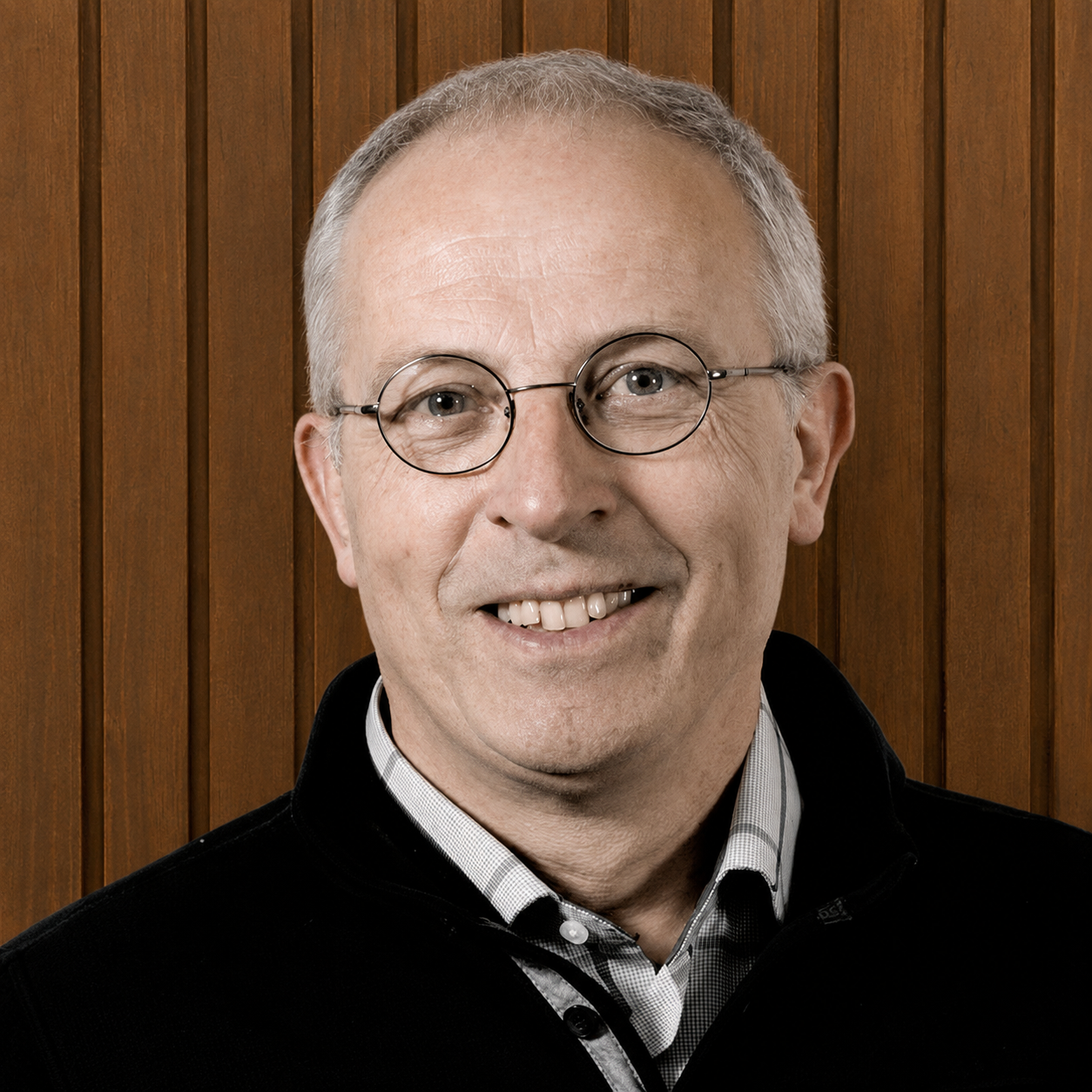 A smiling middle-aged man with glasses, short gray hair, wearing a checkered shirt and black sweater in front of a wooden panel background.