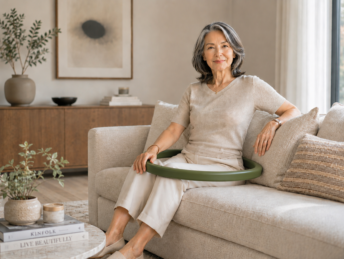 An elderly woman sitting on a cream-colored sofa with a green mobility aid, smiling at the camera in a living room with warm decor.