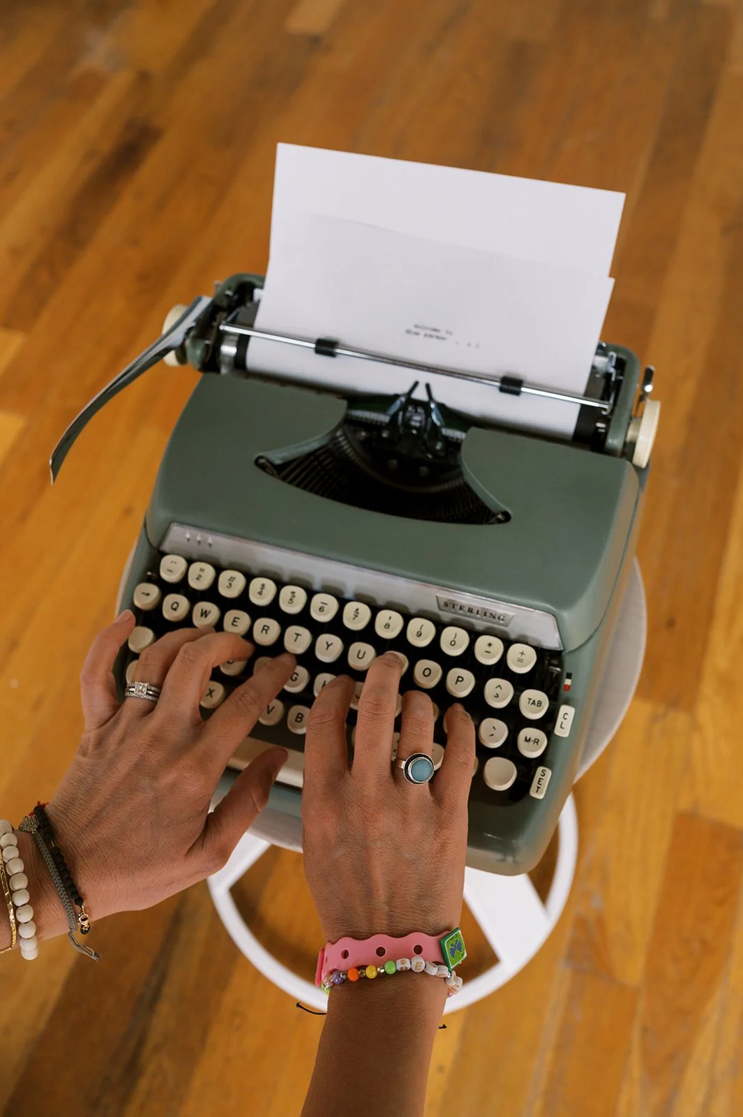A person types on a vintage green typewriter with a sheet of paper loaded, viewed from above, on a wooden floor.