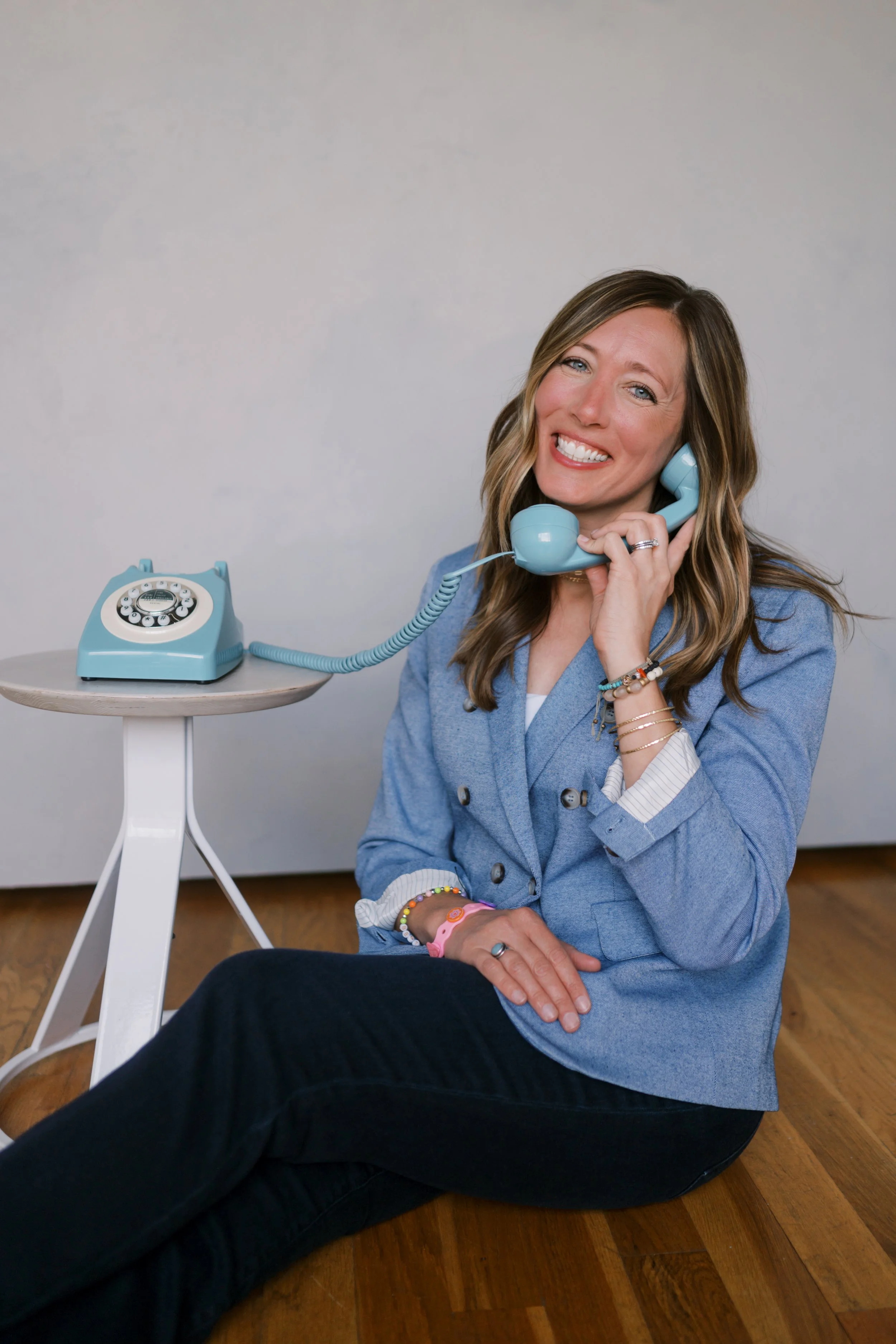 Woman sitting on the floor smiling while talking on a vintage rotary phone, with a retro style matching phone on a small white table