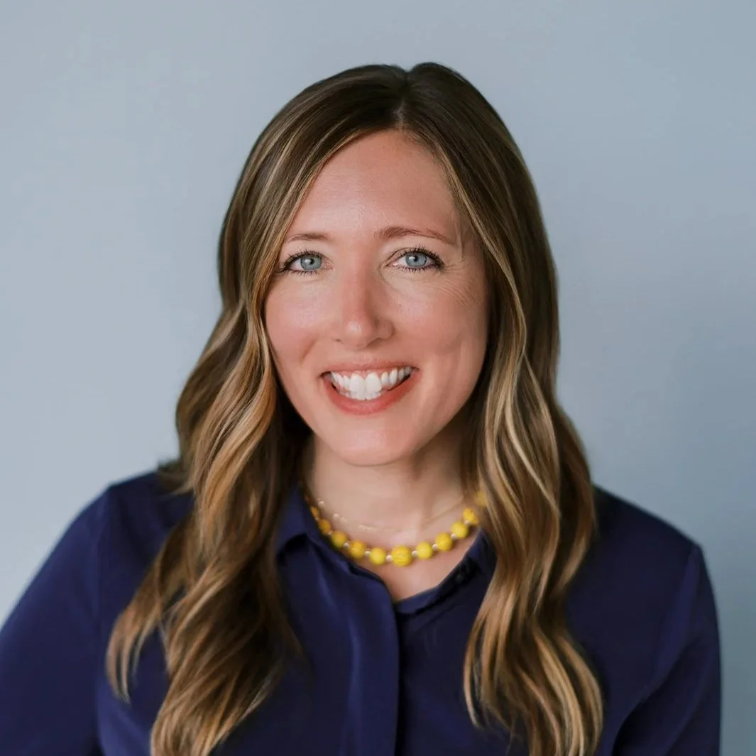 A woman with long wavy light brown hair and blue eyes, wearing a blue shirt and yellow beaded necklace, smiling against a plain light gray background.