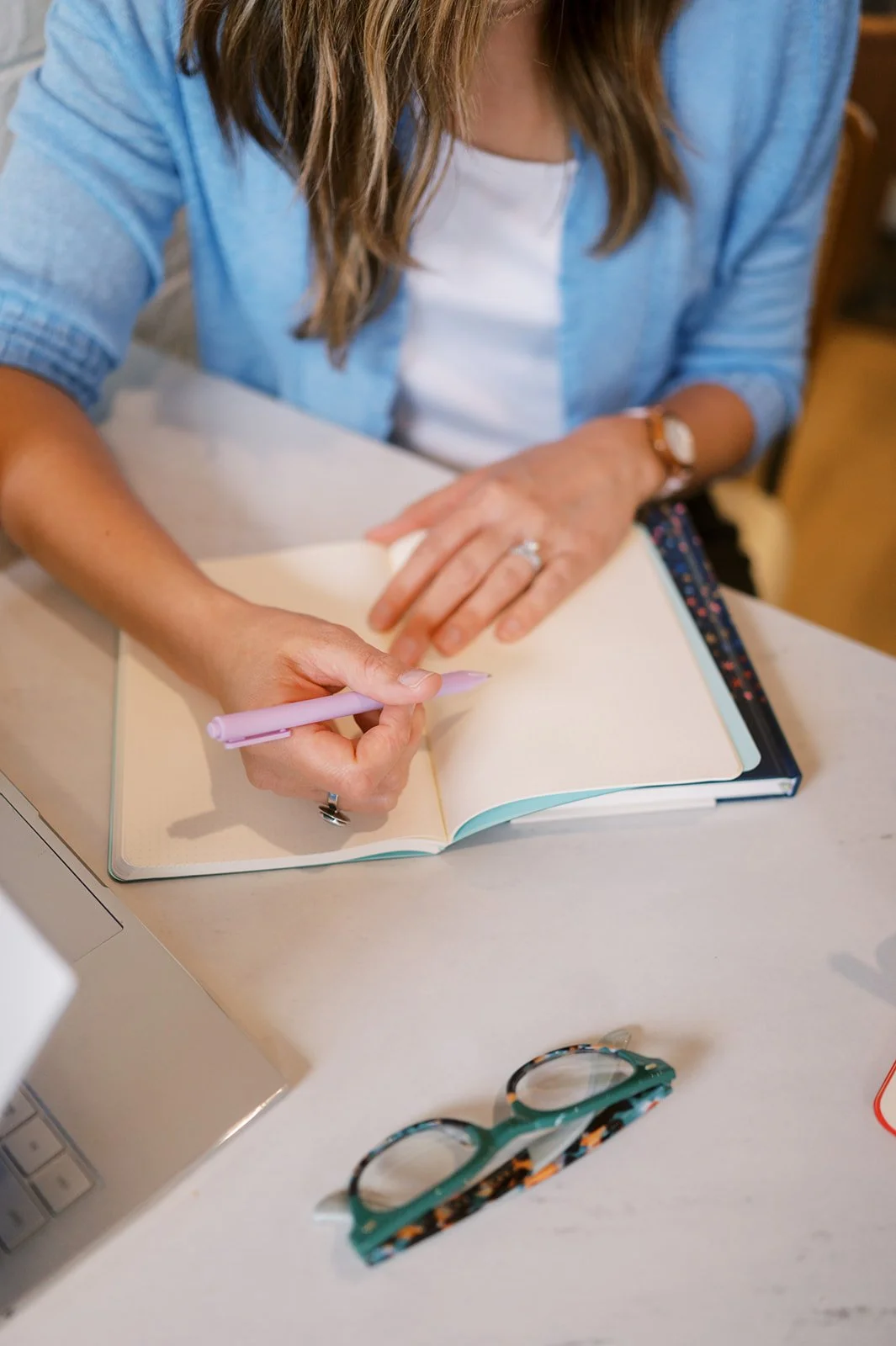 Person writing in a notebook with a pink pen, wearing a blue shirt and a watch, sitting at a desk.