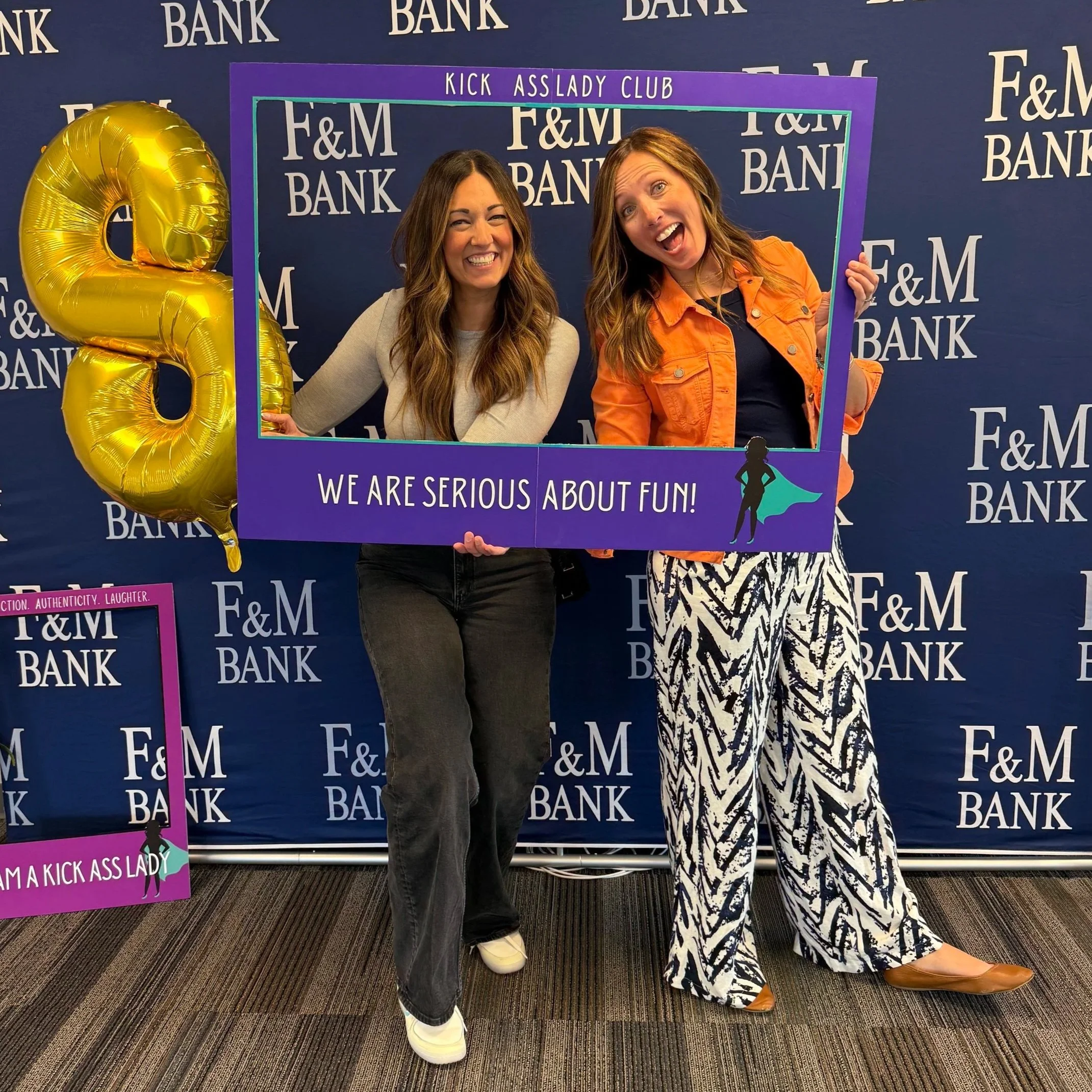 Two women are smiling and posing behind a purple frame with the words "Kick Ass Lady Club" and the slogan "We Are Serious About Fun!" written on it. One woman in a light sweater and jeans holds a large, gold, balloon letter 'Q'. The background features a dark blue backdrop with the repeated logo "F&M Bank."