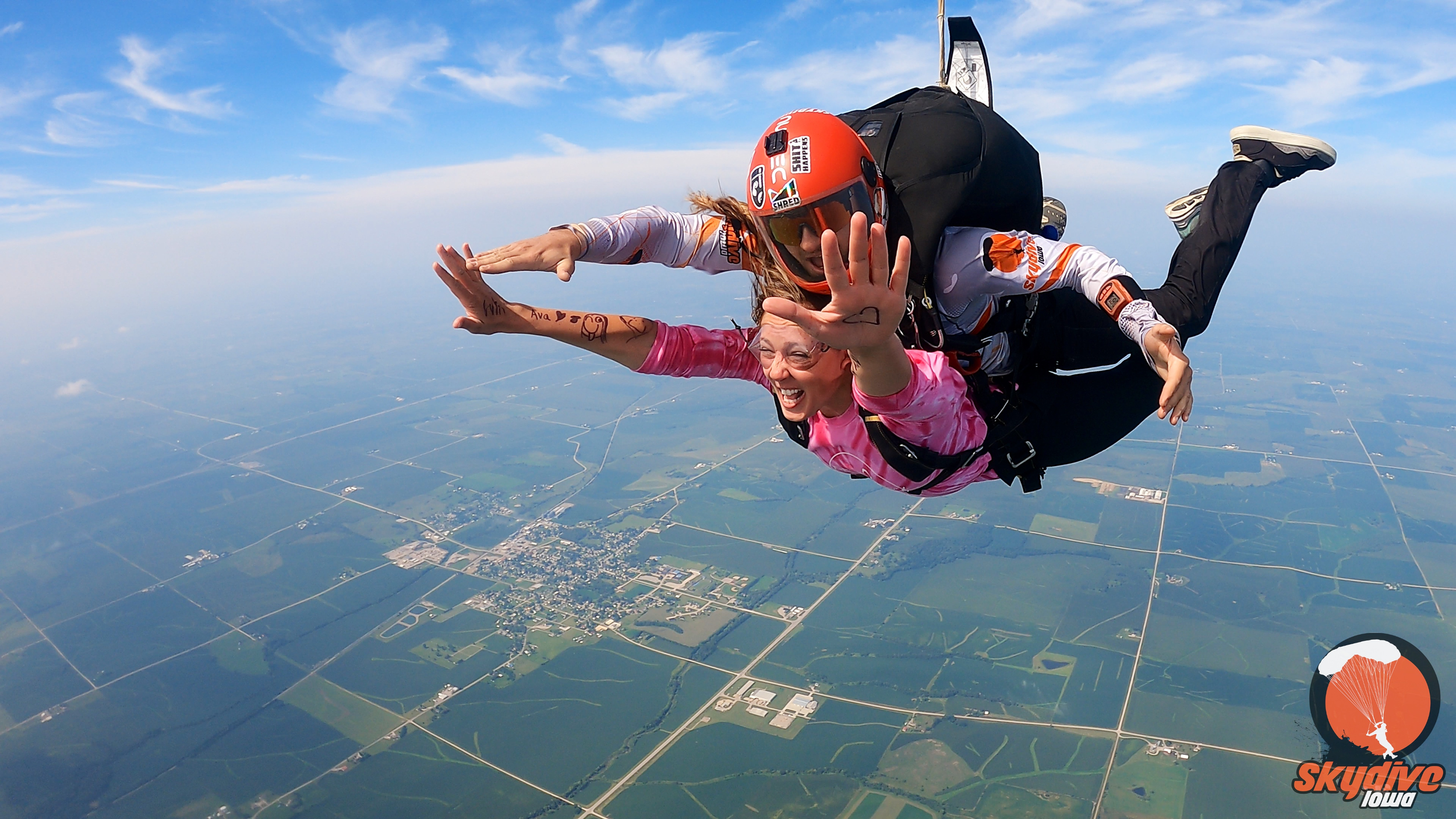 Two women skydiving in a tandem jump over a green landscape with roads and fields, wearing helmets and jumpsuits, smiling and making gestures.