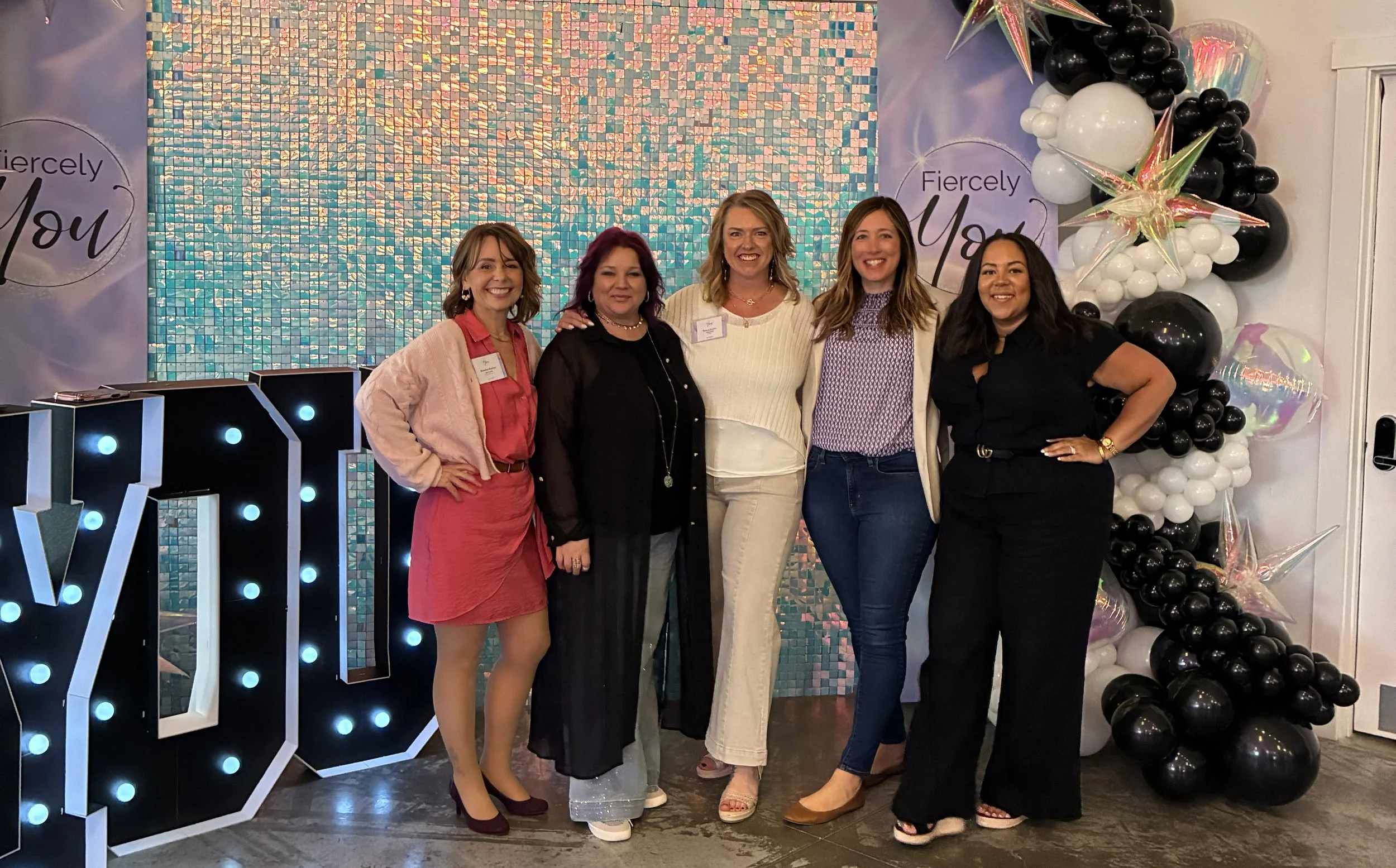 Five women standing and smiling with a shiny background