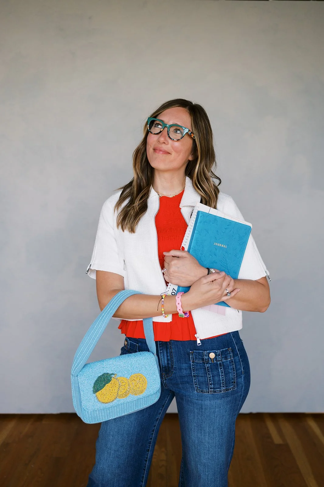 A woman with glasses, wearing a white short-sleeved jacket over a red top, holding a blue notebook, with a blue pineapple-themed bag over her shoulder, standing against a plain background.