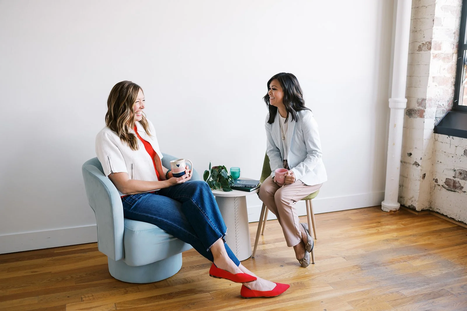 Two women sitting and talking in a bright, minimalist room with wooden floors, holding mugs, and smiling at each other.