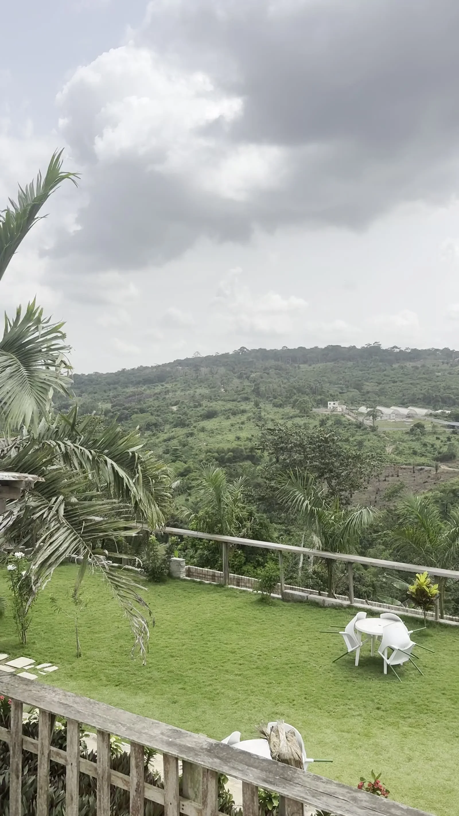 A backyard with a mountain view and cloudy sky, featuring a well-maintained green lawn, outdoor chairs and tables, and tropical plants along the fence.
