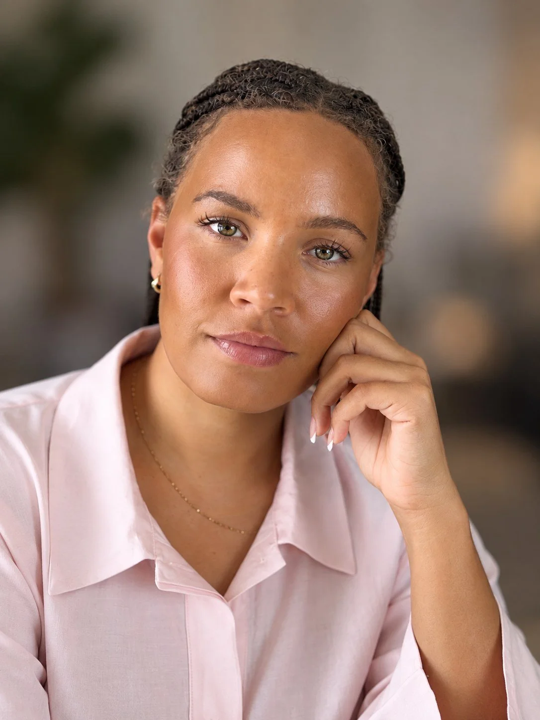 A woman with braided hair and hoop earrings resting her chin on her hand, wearing a light pink shirt and a delicate necklace.