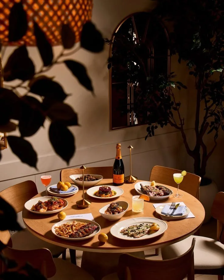 A round wooden dining table set with various dishes, including a bottle of champagne, cocktails, and plated food. There are four chairs around the table and a mirror on the wall reflecting some of the dinnerware and food. The setting is indoors with warm lighting and decorative plants.