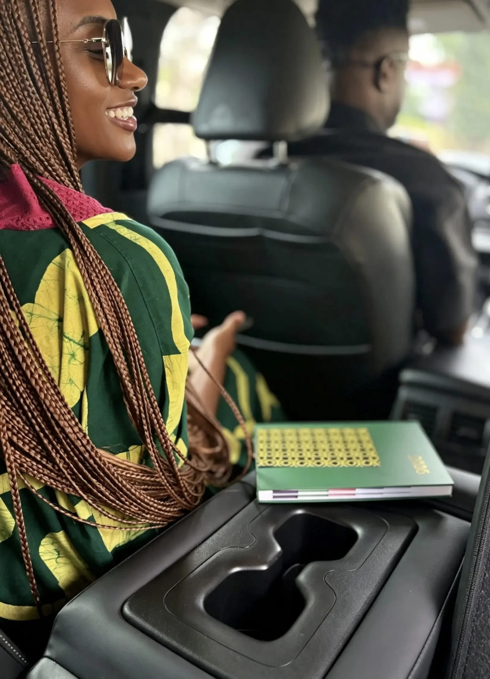 A woman with braided hair and sunglasses sitting in a car, smiling, with a green book or magazine on her lap and a man driving in the background.