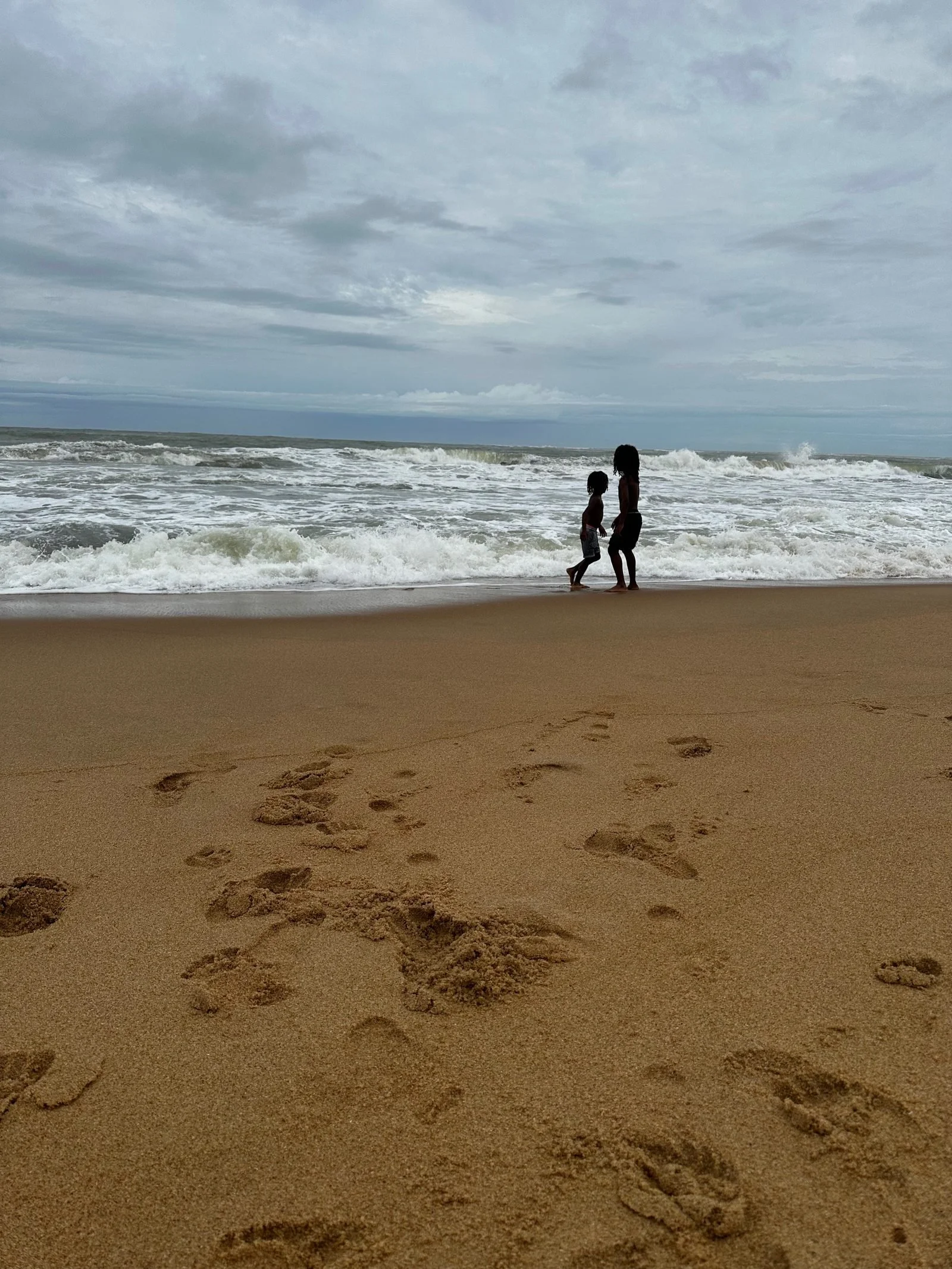 Two children walking on a sandy beach with footprints in the foreground, ocean waves, and a cloudy sky in the background.