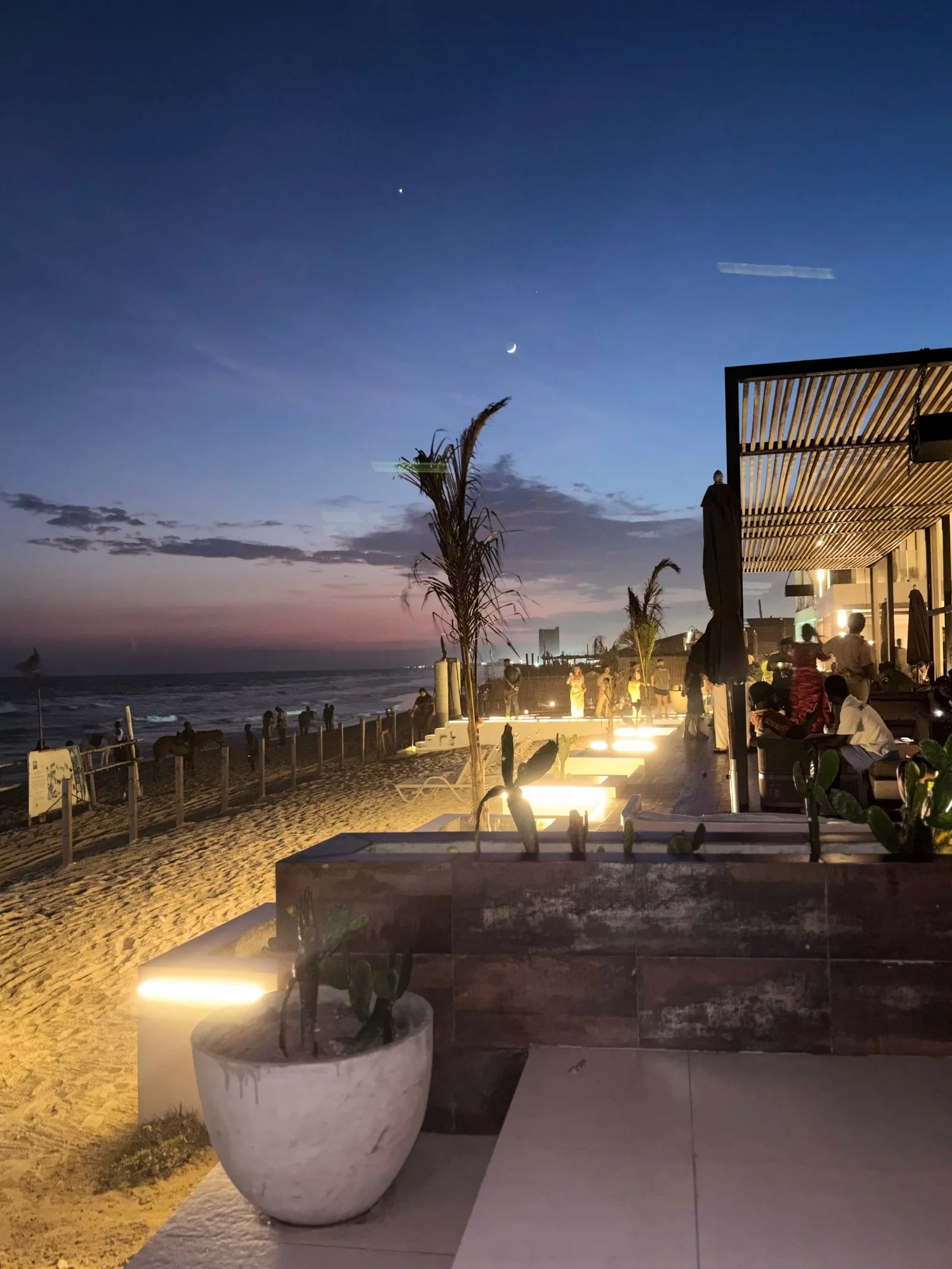 Beachside restaurant or bar during sunset with people dining and relaxing, palm trees, sand, and ocean in the background, illuminated lighting creating a warm ambiance.