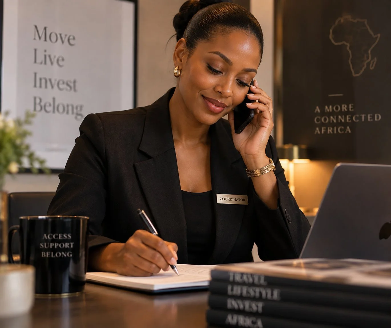 A woman with a dark bun hairstyle, wearing a black blazer with a 'Coordinator' name tag, sitting at a desk with an open laptop, talking on her phone, and taking notes with a pen. The background features motivational and regional posters.