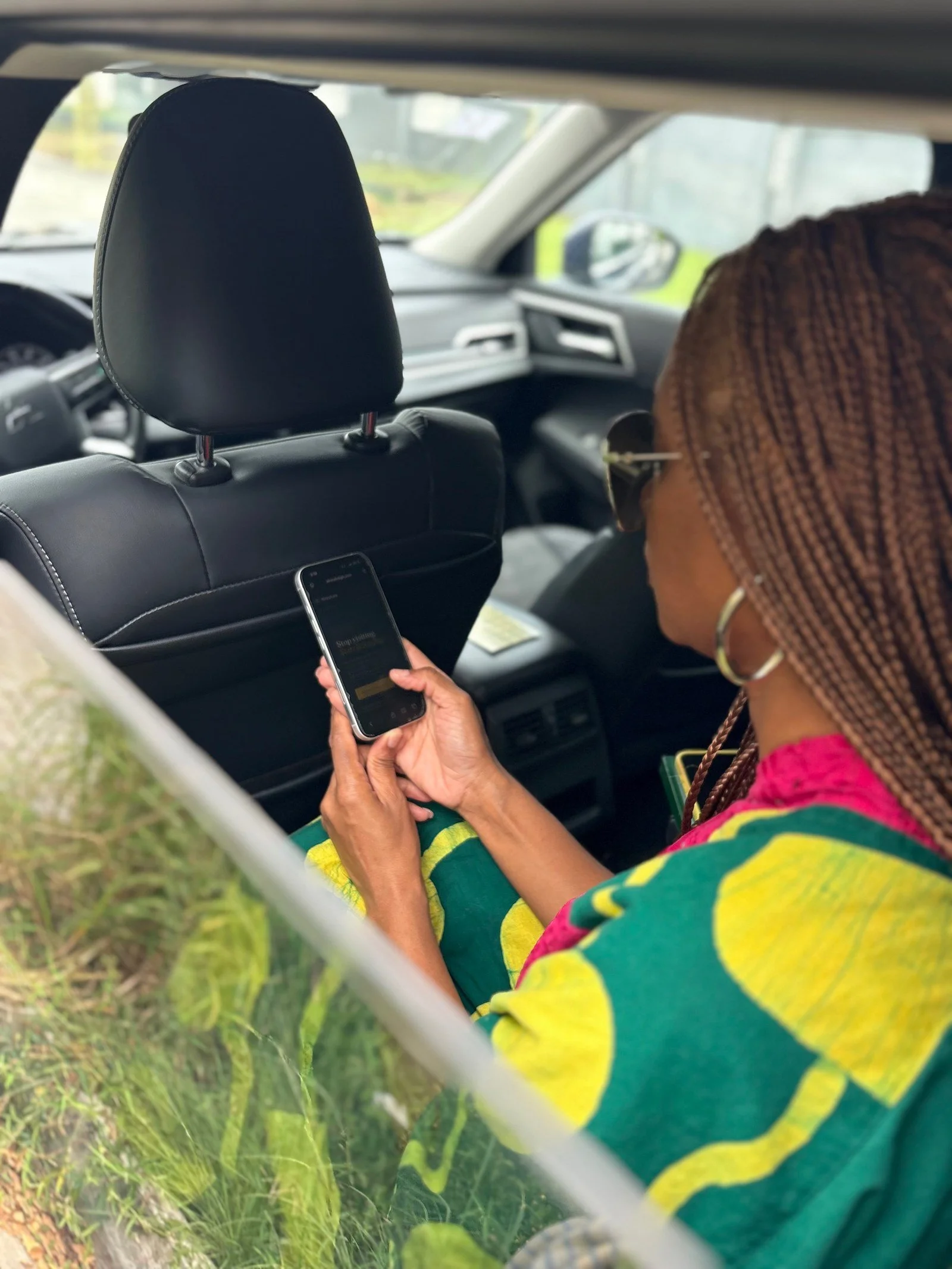 Person sitting in the backseat of a vehicle, looking at their phone, wearing sunglasses and a colorful striped shirt with braids in their hair.
