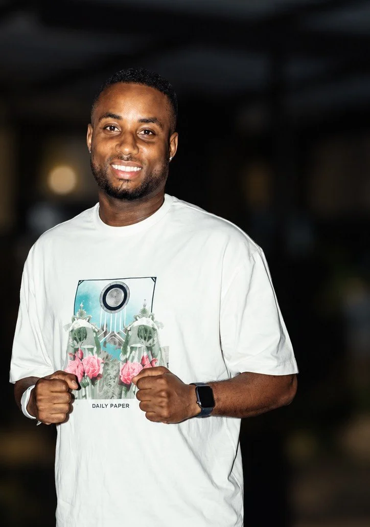 Man smiling, wearing a white t-shirt with a graphic of flowers and a cassette tape, and a smartwatch, standing indoors.