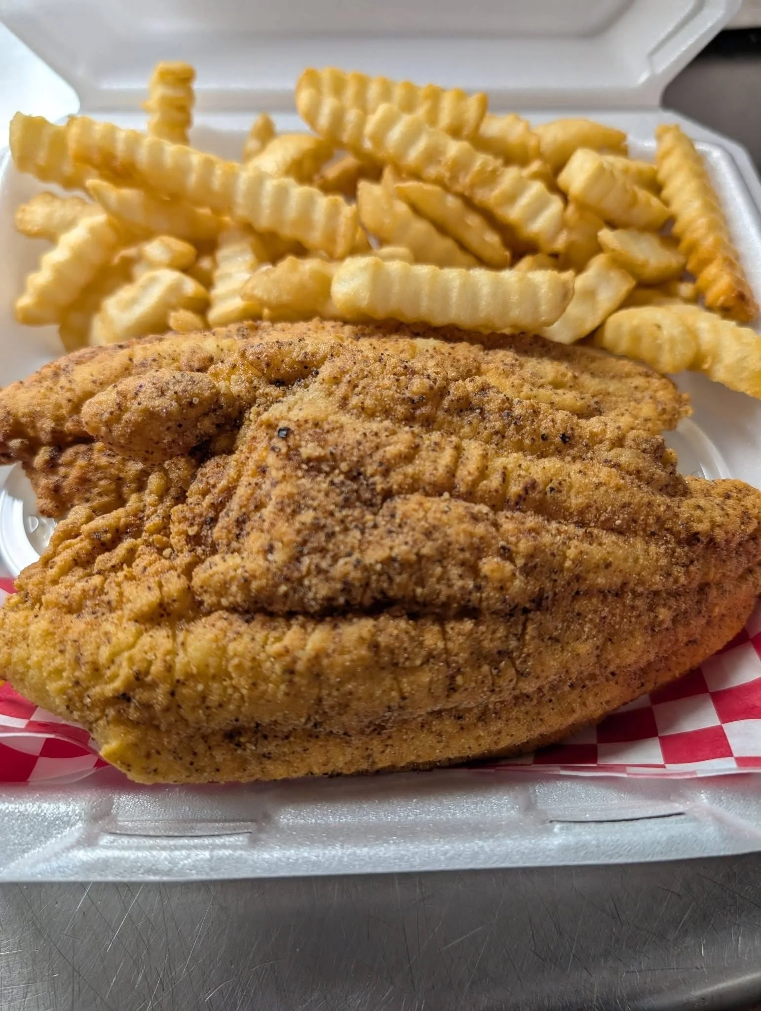Close-up of a fried chicken tender and crinkle-cut French fries in a takeout styrofoam container lined with red-and-white checkered paper.