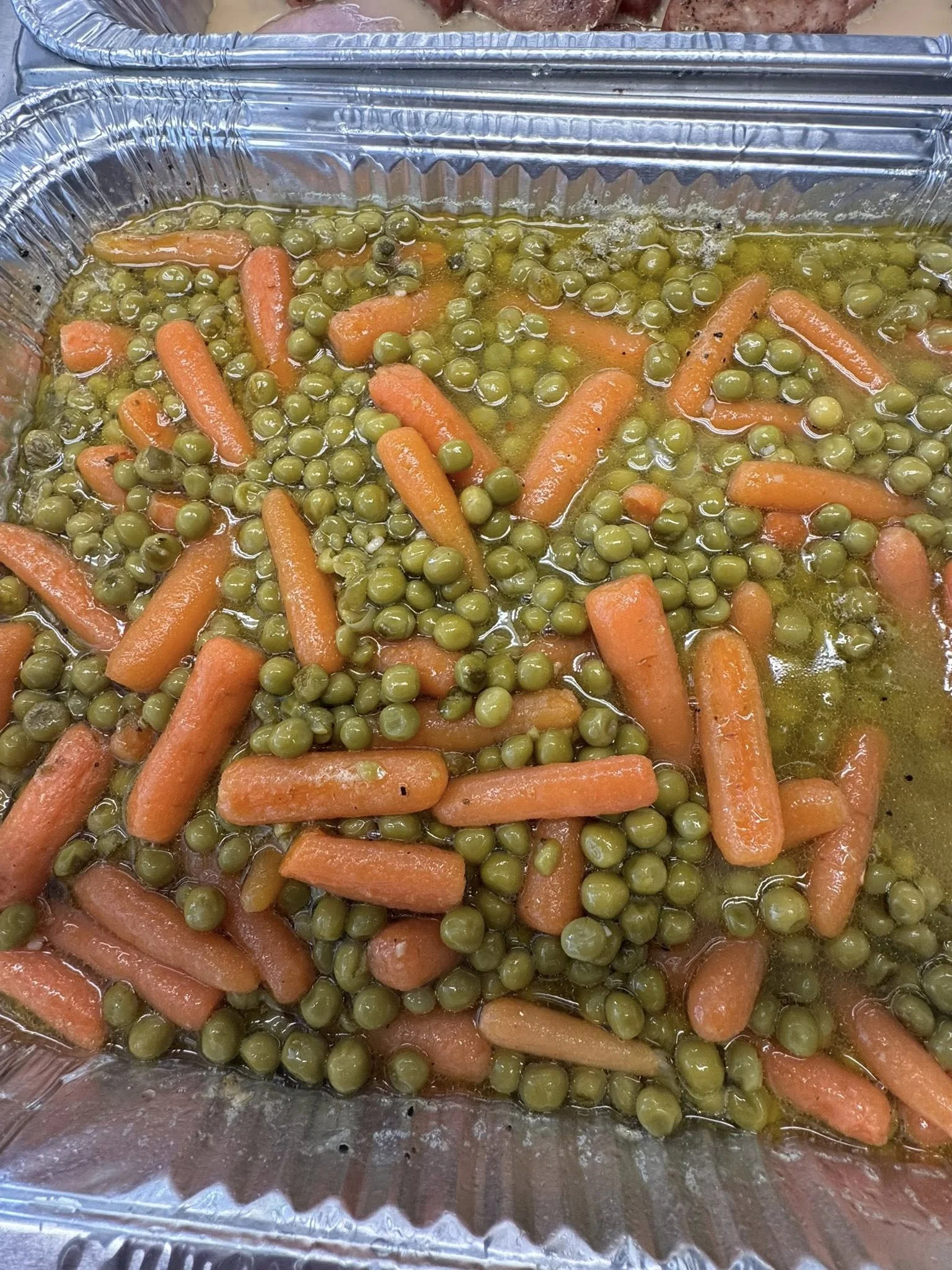 Cooked carrots and green peas in a aluminum tray with some liquid at the bottom.