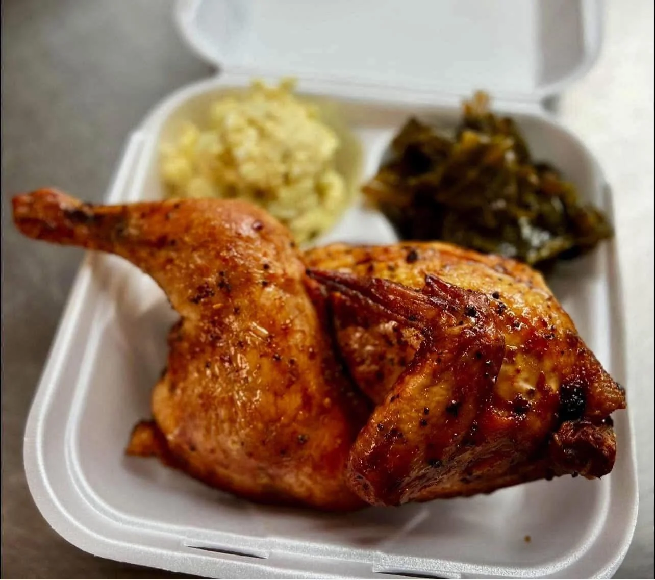 To-go container holding a piece of baked chicken, macaroni and cheese, and collard greens.