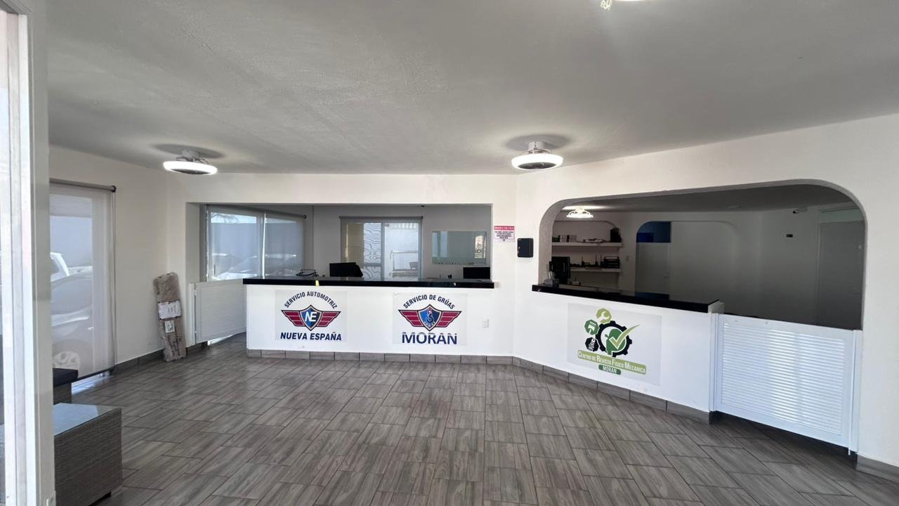 Recepción de un taller mecánico con dos logotipos en la mesa, uno de 'Nuevo España' y otro de 'Morán', y un cartel en la pared.