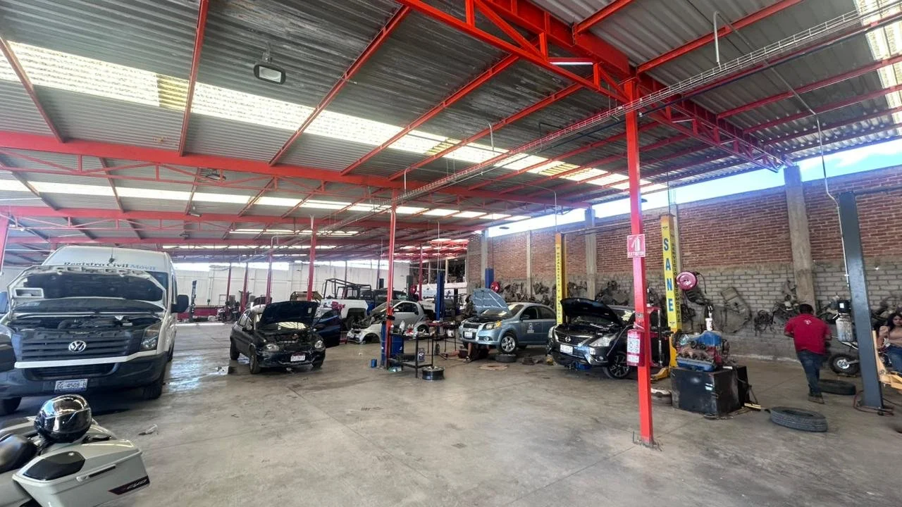 Interior de un taller mecánico con varios autos en reparación y personas trabajando, con estructura de techos metálicos y pared de ladrillos.