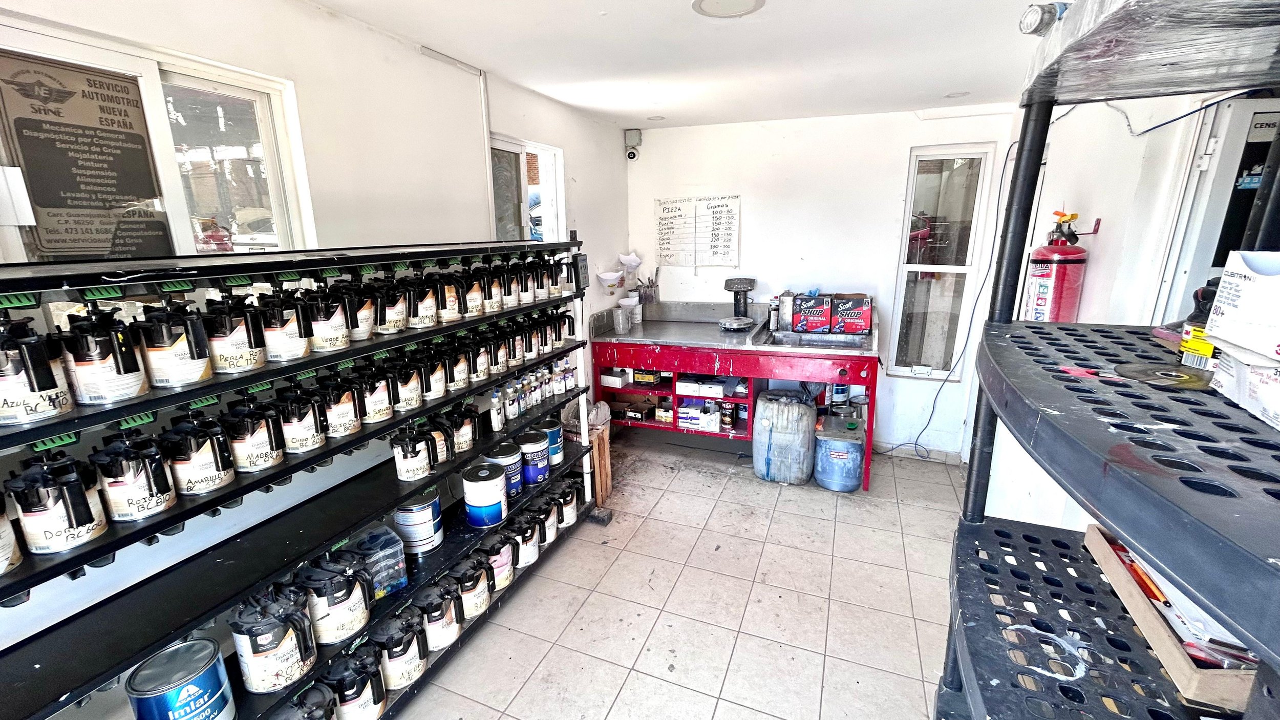Interior de un taller de reparación de automóviles con estantes llenos de pinturas en envases, una mesa de trabajo, y equipos de herramientas.
