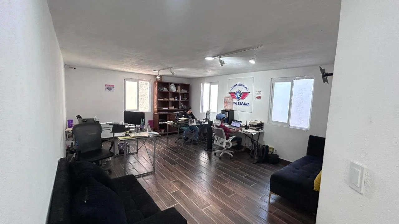 Oficina con varias mesas, sillas y computadoras, y tres personas trabajando en ellas, decorada con un cartel en la pared que dice 'SERVICIO AUTOMOTRIZ NUEVA ESPAÑA'.