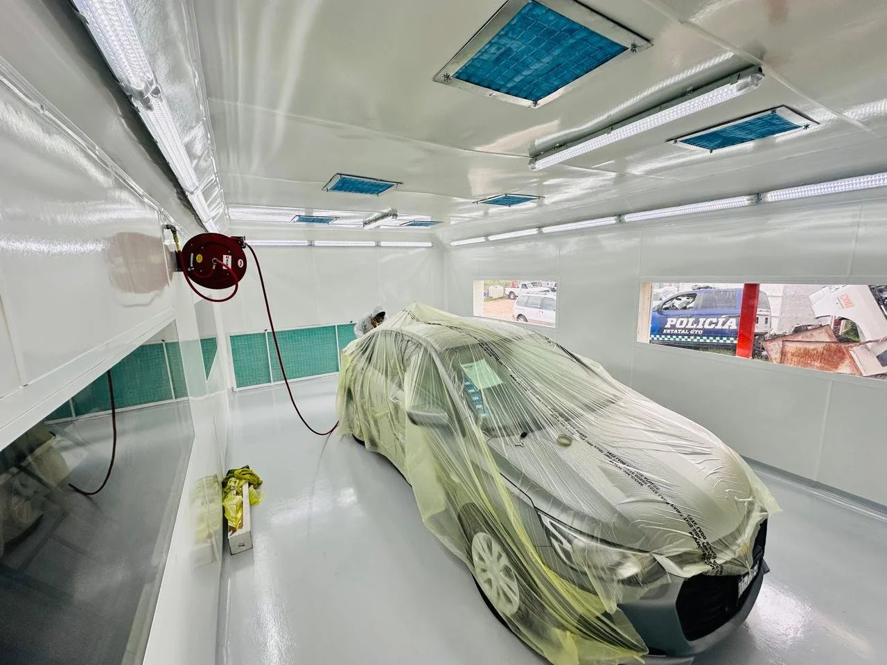 Coche en cabina de pintura, cubierto con plástico protectora, y trabajador en el fondo, en una instalación de pintura de autos.