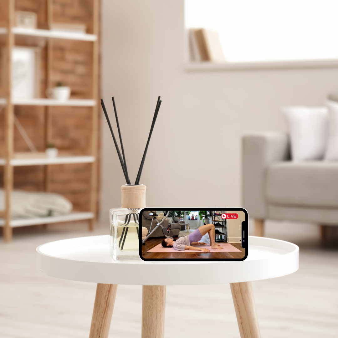 A white round table with a reed diffuser and a smartphone displaying a live yoga class in a bright, modern living room