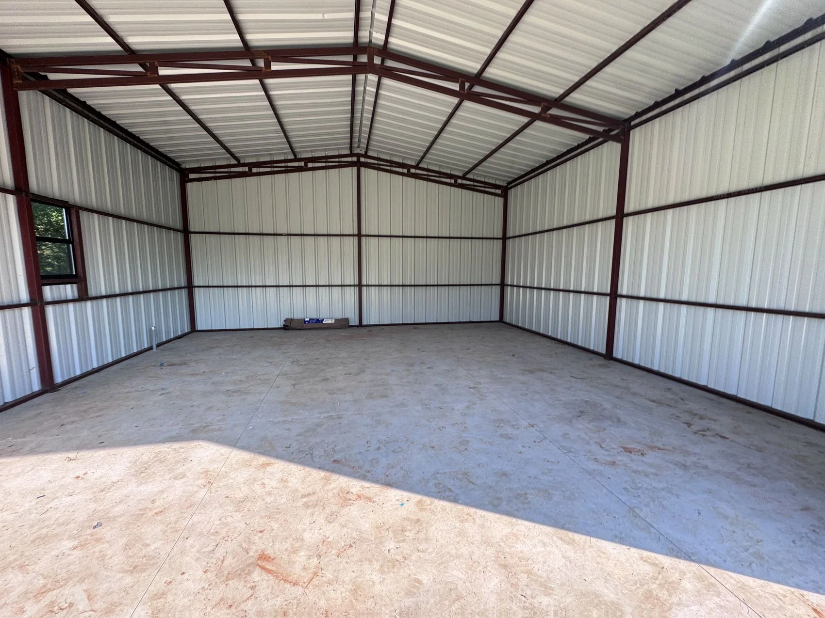 Empty indoor metal storage building with concrete floor, white paneled walls, and a high metal roof. There is a small window on the left wall and a rolled-up item on the back wall.