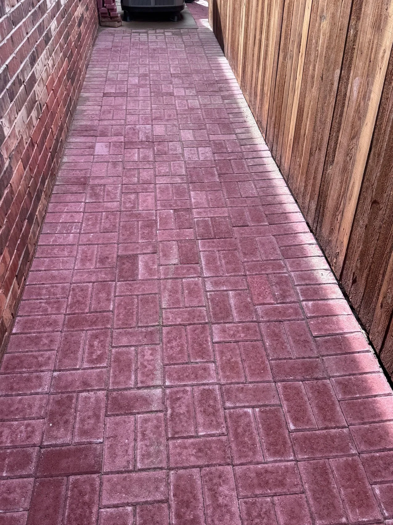 A narrow outdoor pathway with pinkish-red brick paving, flanked by a red brick wall on the left and a wooden fence on the right.