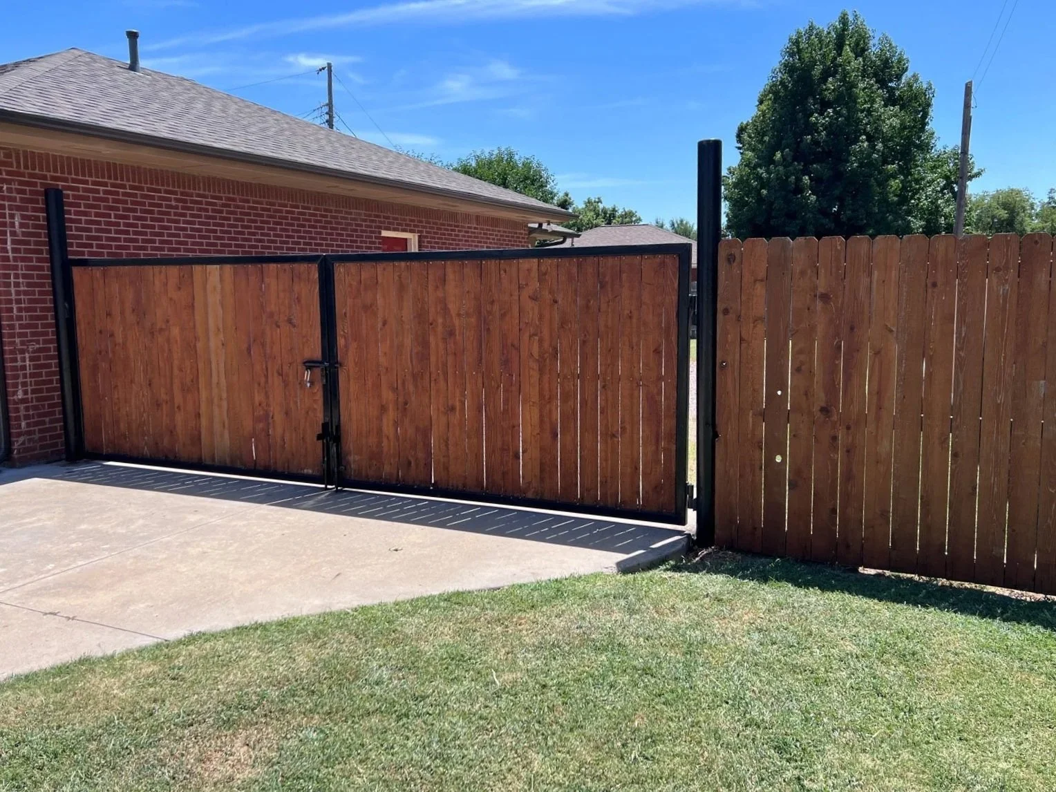 A wooden gate with a lock and metal frame, next to a wooden fence, with a brick house and green trees in the background on a sunny day.