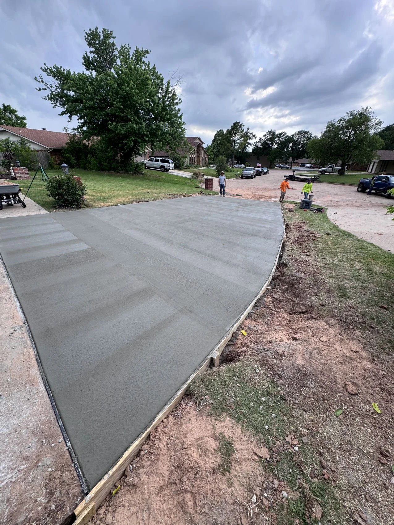 Newly poured concrete driveway with a smooth surface, surrounded by dirt and grass, with construction workers working in the background on a residential street.