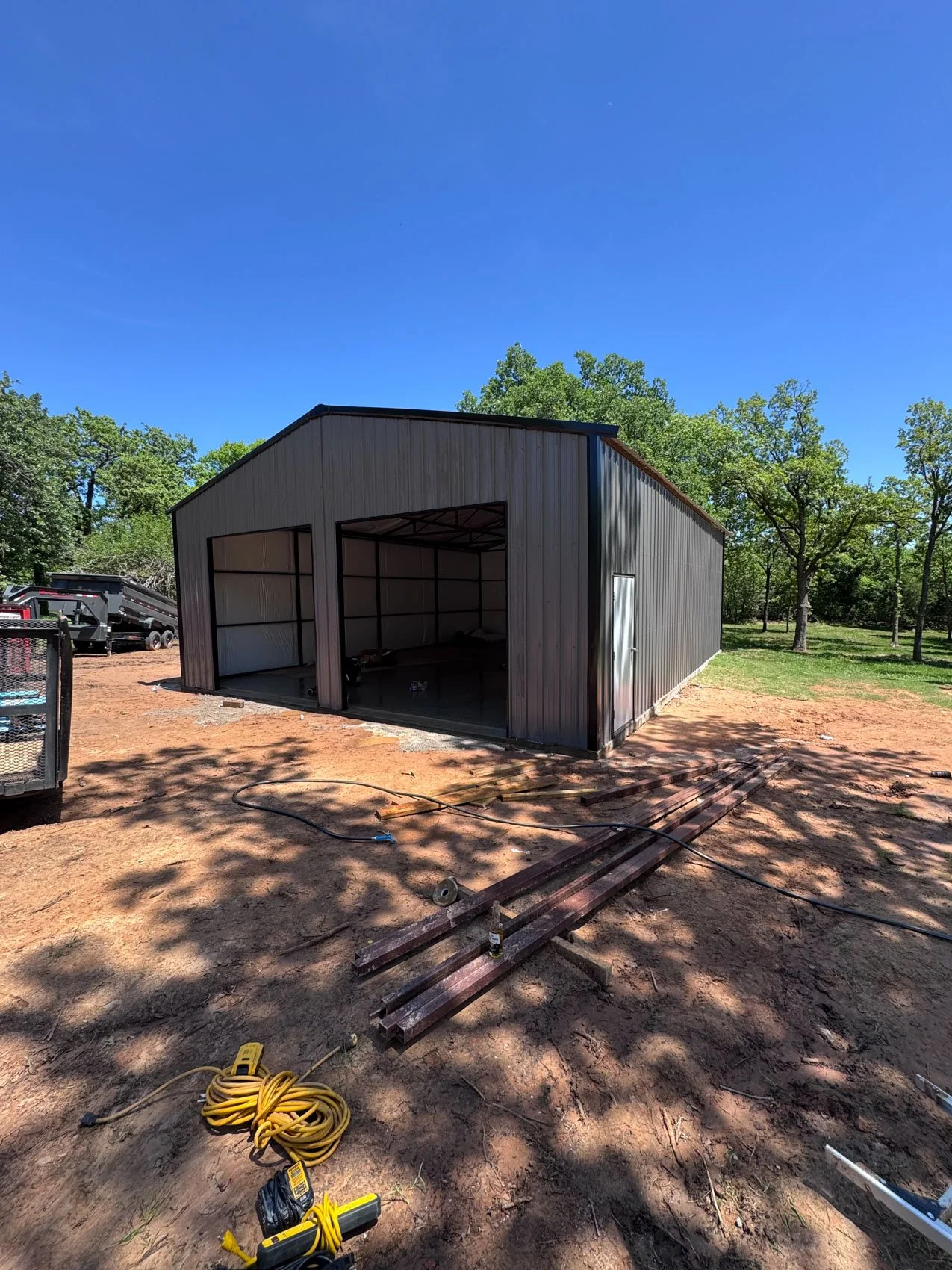 Steel shed under construction on a dirt lot with trees in the background, construction tools and materials on the ground.