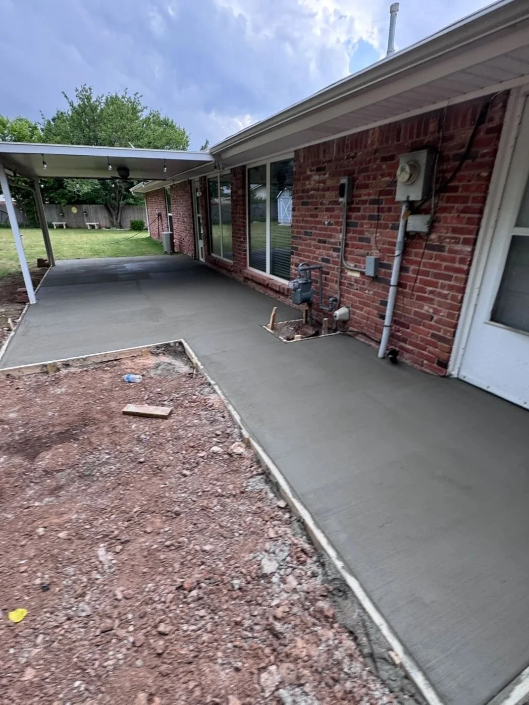 Recently poured concrete patio with a small section of dirt and gravel in the foreground, next to a brick house with sliding glass doors. There's a covered patio area with string lights in the background and a grassy yard with trees.
