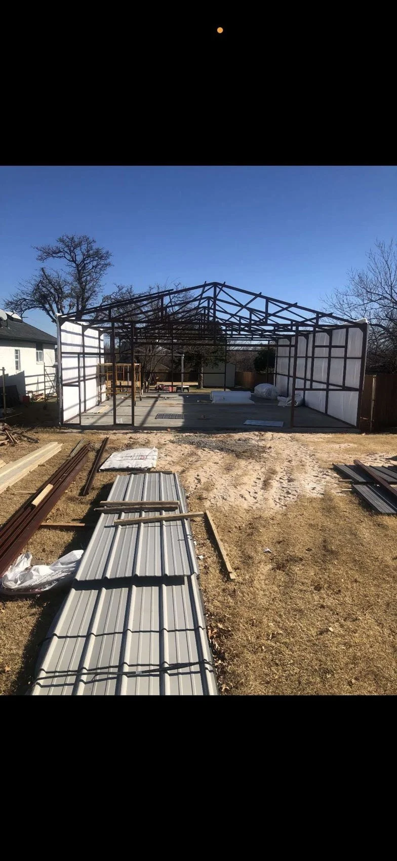 Construction site where a metal frame structure for a building or greenhouse is being assembled, with building materials like metal panels and lumber in the foreground and a clear blue sky above.