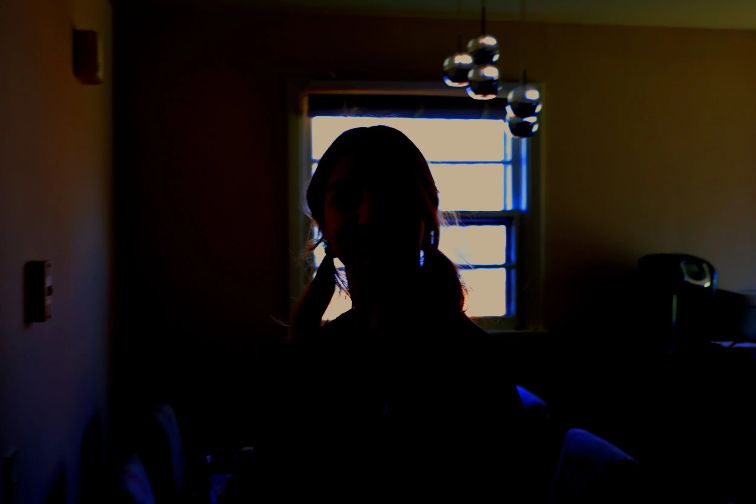 Silhouette of a person with long hair and pigtails standing in front of a window with light streaming in, in a dimly lit room.