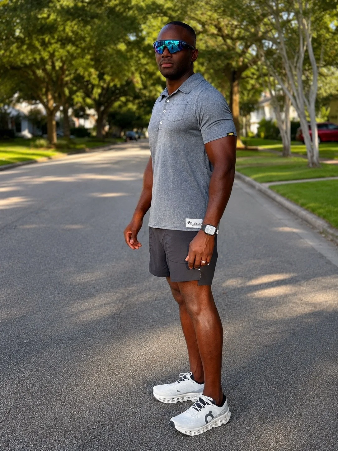 Man wearing sunglasses, gray polo shirt, shorts, and white athletic shoes standing on a tree-lined suburban street