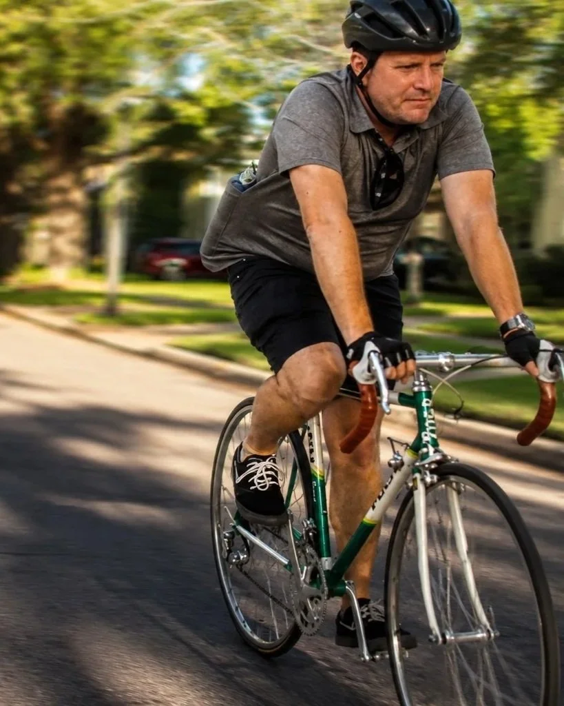 Man wearing a bicycle helmet rides a green road bike through a tree-lined neighborhood street.