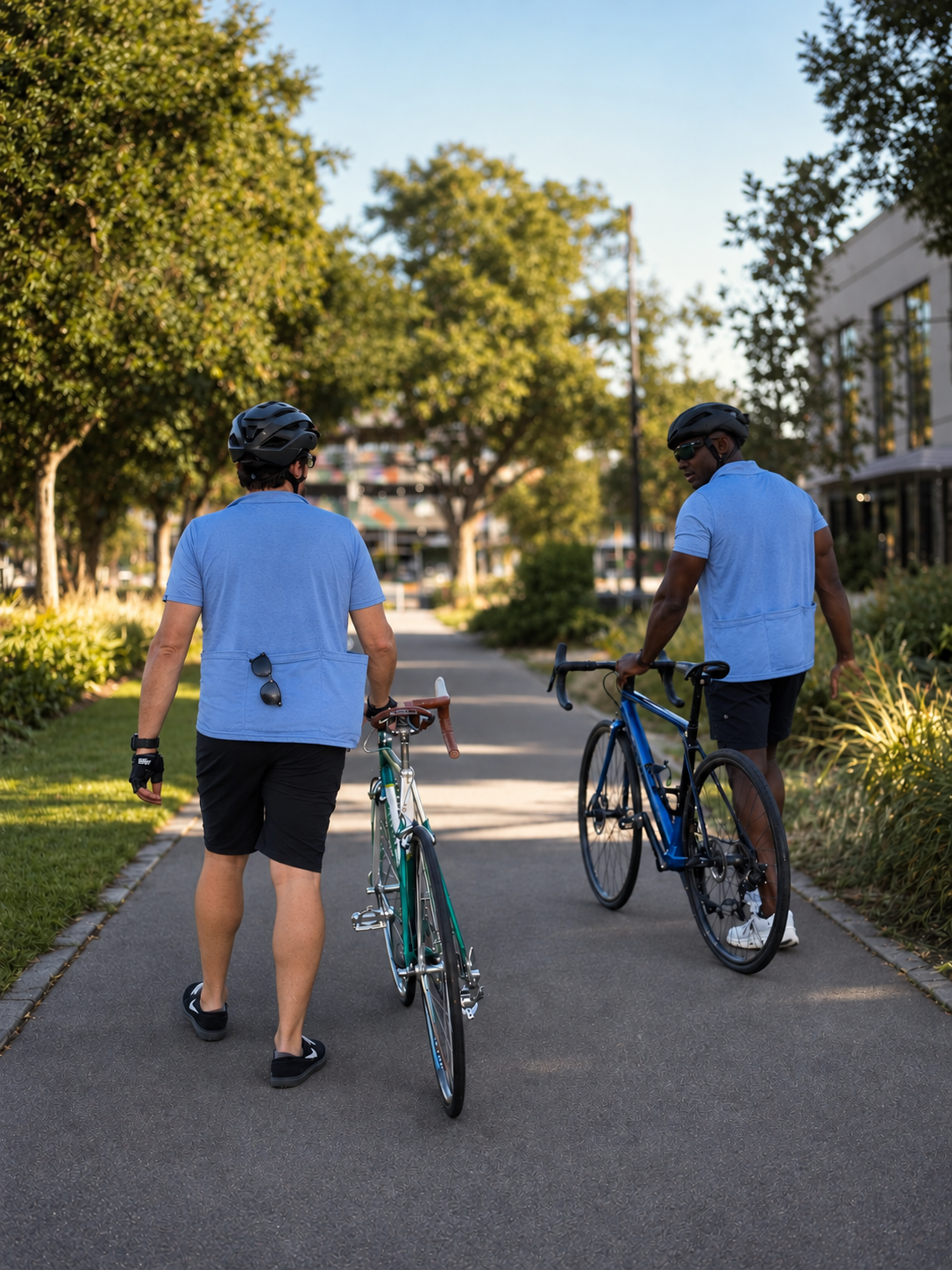 Two cyclists wearing helmets walk their road bikes along a tree-lined urban path.