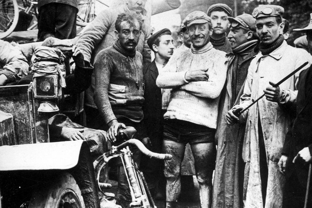 Group of men, some wearing goggles, around an old vehicle and a bicycle, outdoors.