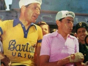 Two men, one in a yellow and blue cycling jersey and the other in a pink polo shirt with a white cap, are standing outdoors with a crowd. The man in the yellow jersey appears to be emotional or crying. The man in pink is holding something in his hands.