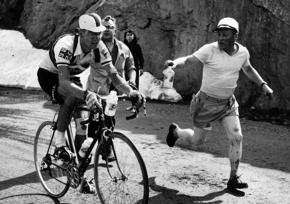 A man on a bicycle with a water bottle, pedals on a mountain road. Another man running alongside offers a water bottle, with two other individuals following behind. The scene appears to be during a cycling event in a mountainous area.