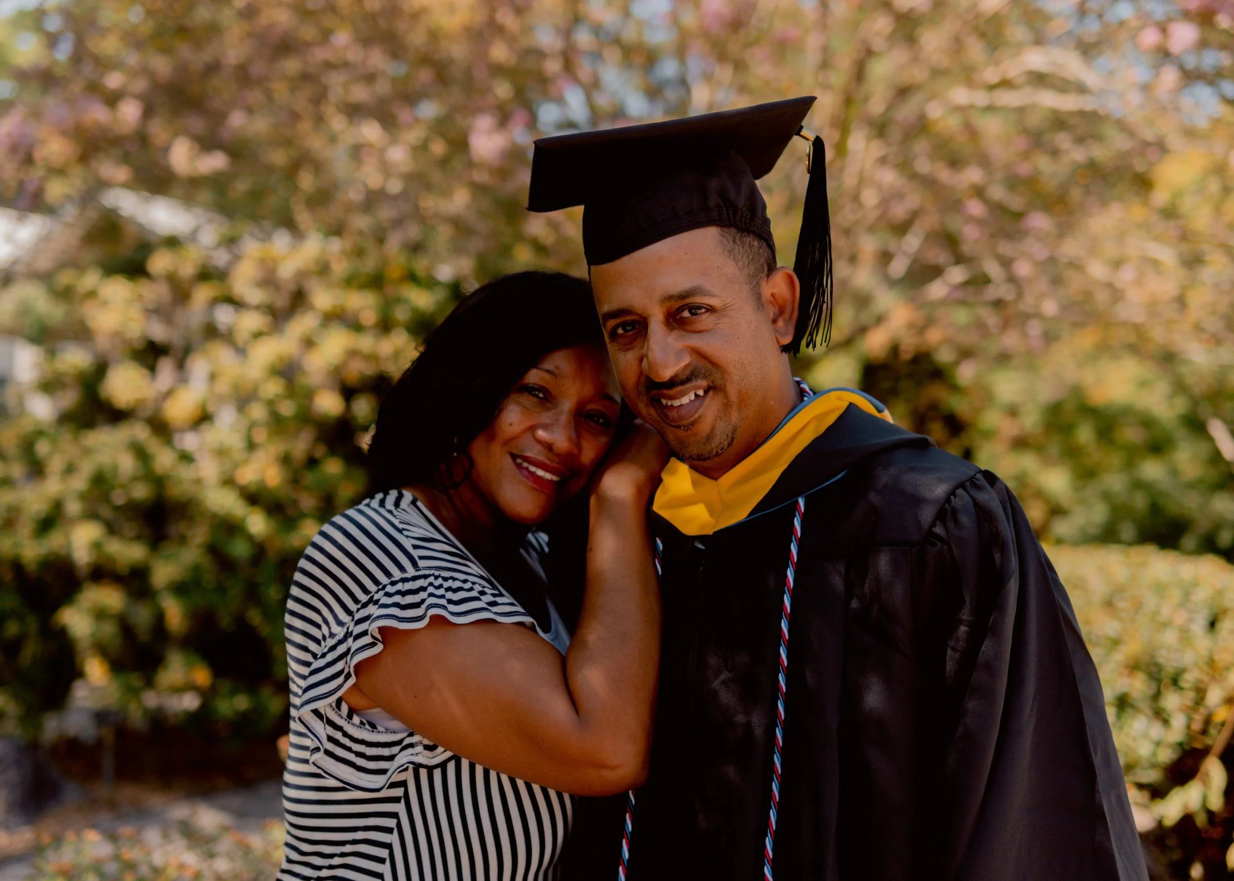 A woman and a man in a graduation gown and cap embrace outdoors with trees displaying fall foliage in the background.